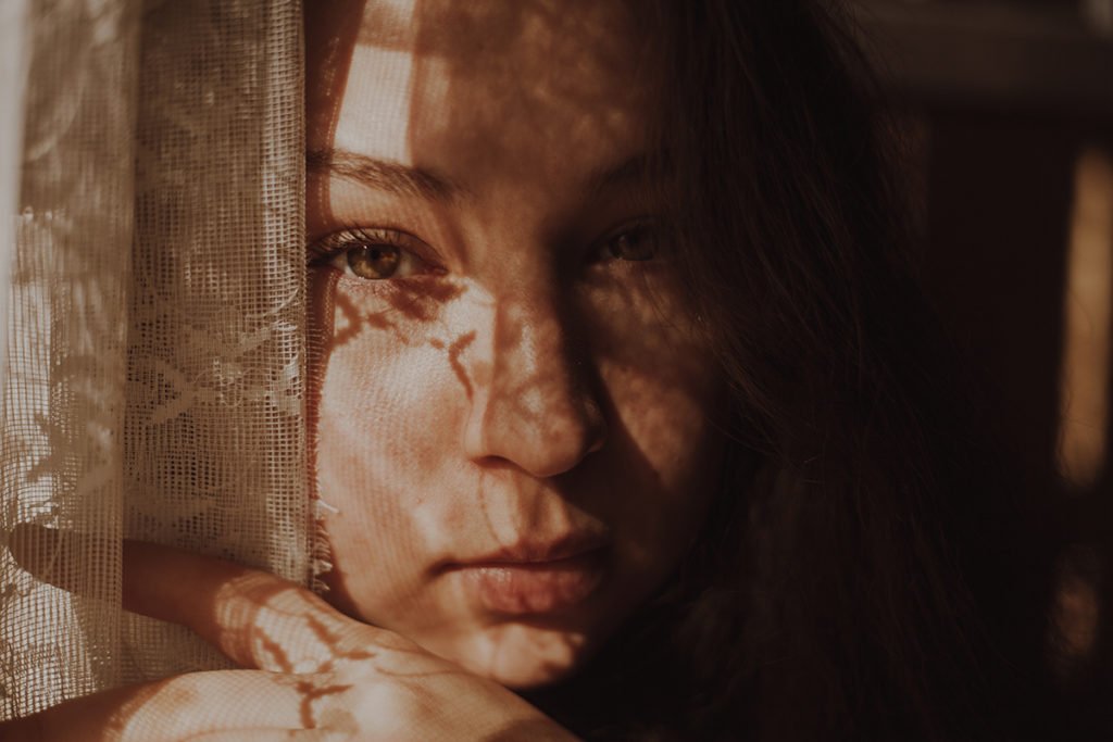 Portrait of a girl’s face holding a curtain. Her face is covered in shadows.