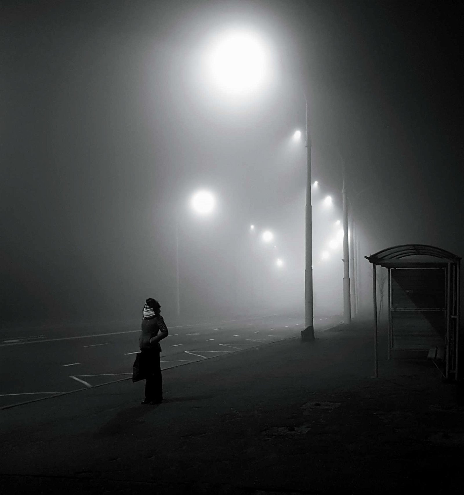 black and white image of woman standing next to bus stop on a foggy evening.