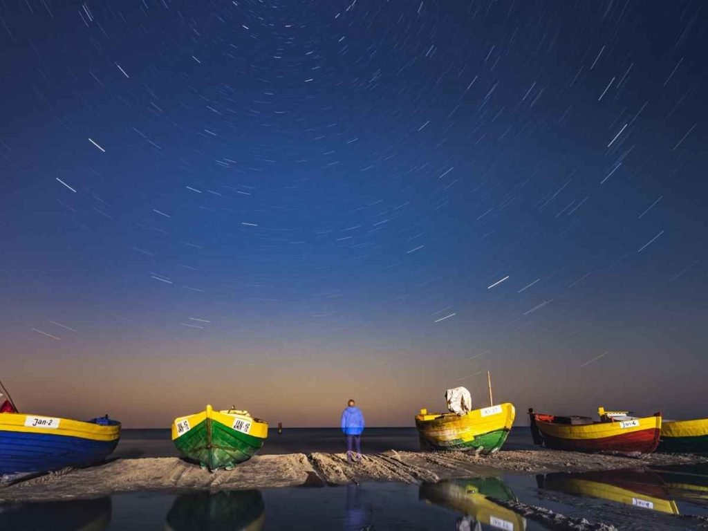 person standing on the shore next to boats looking at stars in the night sky.